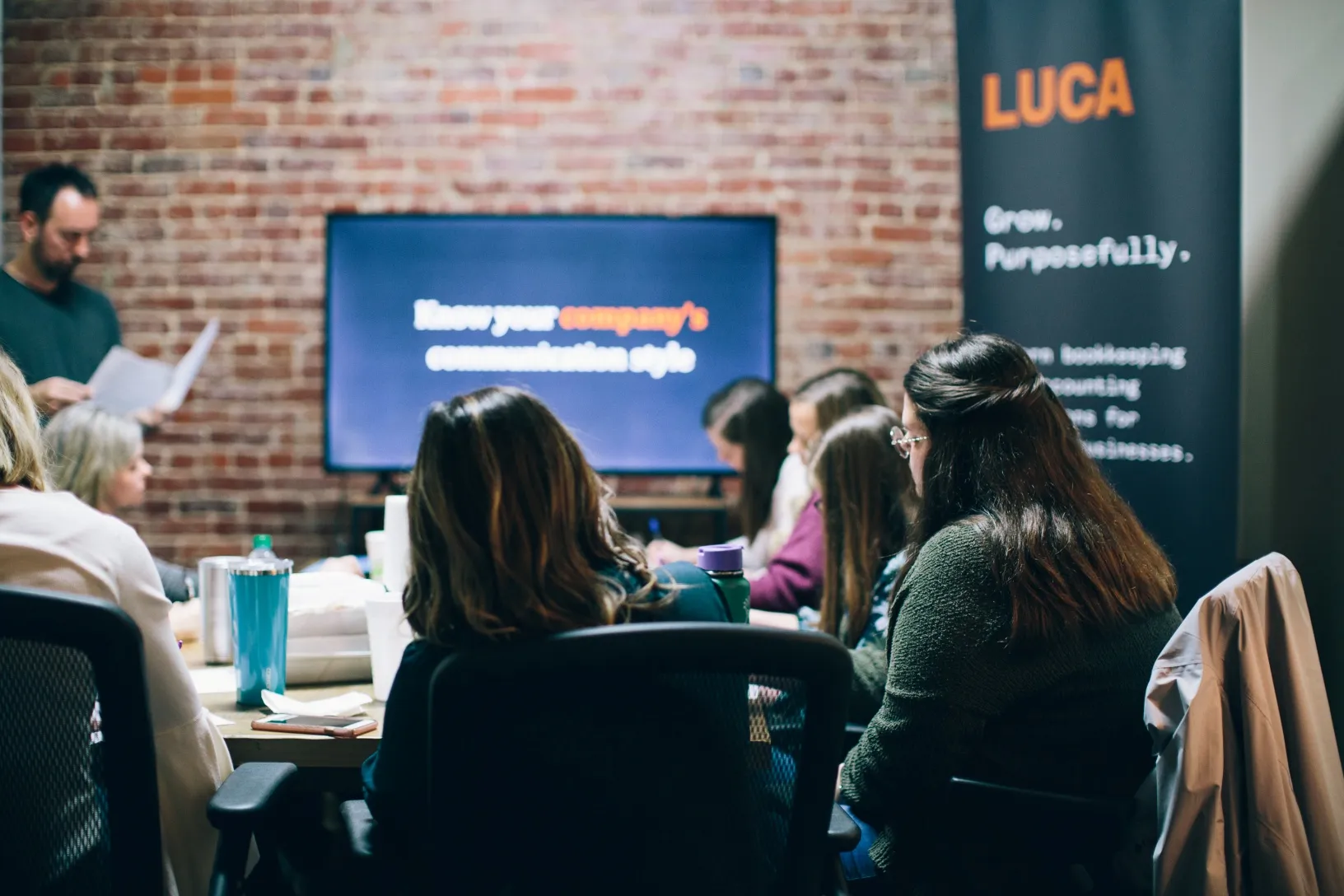 LUCA team members around a conference table in a brick-walled room, watching a presentation on a wall screen beside a LUCA banner